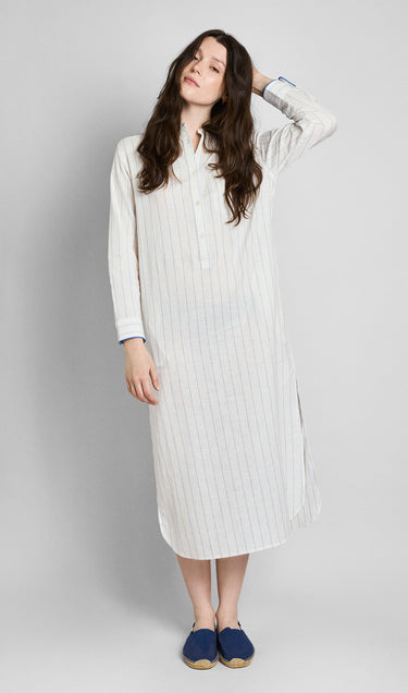A woman wearing a long, white linen button-down kaftan with blue stripes and blue piping details on a plain grey background.