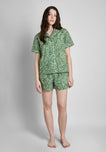 A woman wearing a short sleeve and shorts green pajama set with a white botanical print stands in front of a plain background.