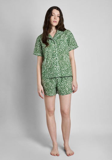 A woman wearing a short sleeve and shorts green pajama set with a white botanical print stands in front of a plain background.
