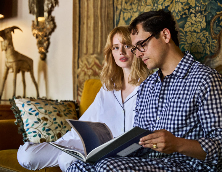 Man and woman sitting together, wearing pajamas (woman in white pajamas, man in navy and white gingham) looking at a book in a decorated room.