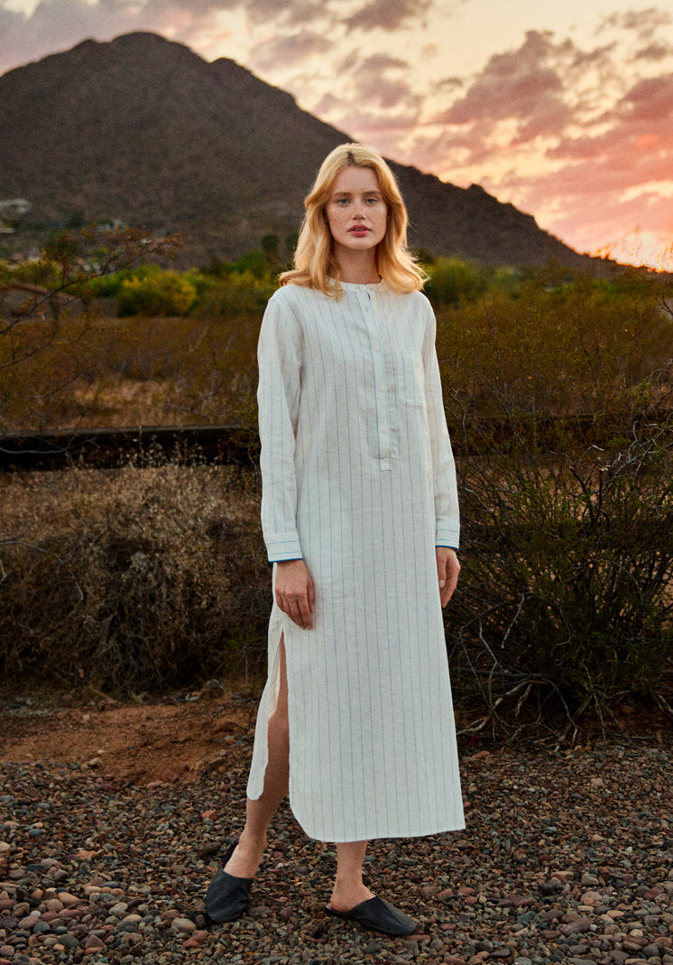 Woman in a long, white linen dress with blue stripes standing in a desert landscape with mountains and sunset.