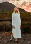 Woman in a long, white linen dress with blue stripes standing in a desert landscape with mountains and sunset.