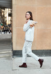 A woman wearing grey and white gingham pajamas walks outside carrying a plastic bag of bagels. 