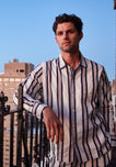 A man wearing white pajamas with navy and red stripes stands on a balcony in front of a blue sky and buildlings.