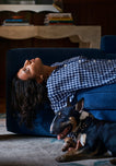 A woman wearing a navy and white gingham pajama top lays on a sofa with her head and hair dangling off the side. A dog lays on the floor next to the sofa.