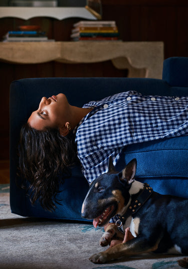 A woman wearing a navy and white gingham pajama top lays on a sofa with her head and hair dangling off the side. A dog lays on the floor next to the sofa.